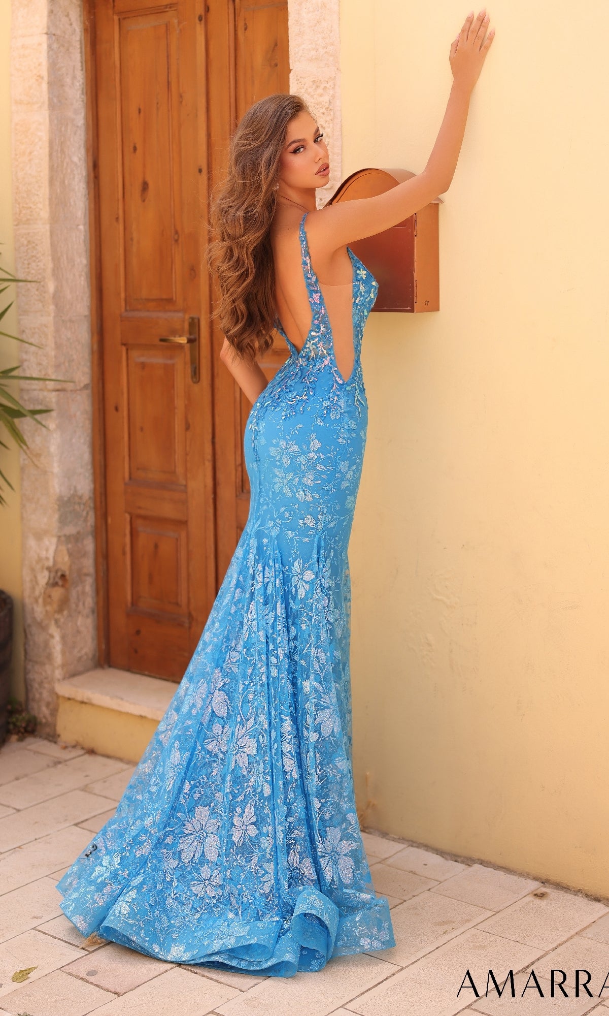 A woman in the Amarra 88275 Sheer-Sides Long Glitter Prom Dress, featuring a fitted mermaid silhouette and low back, stands outdoors by a wooden door and yellow wall, elegantly glancing over her shoulder.